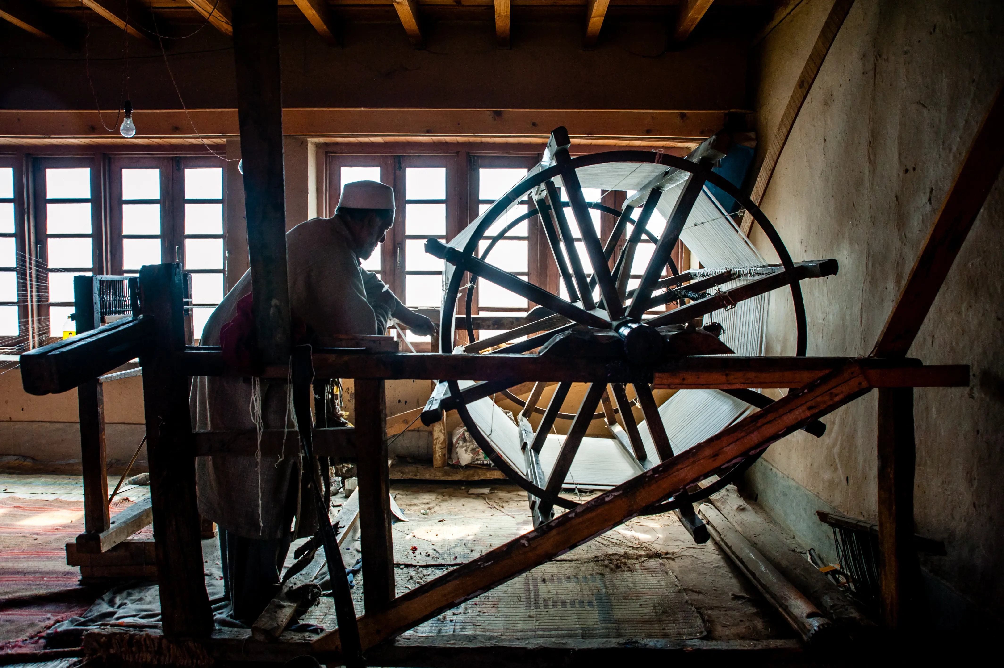 A silhouetted craftsman working at a large wooden warping wheel, backlit by a window