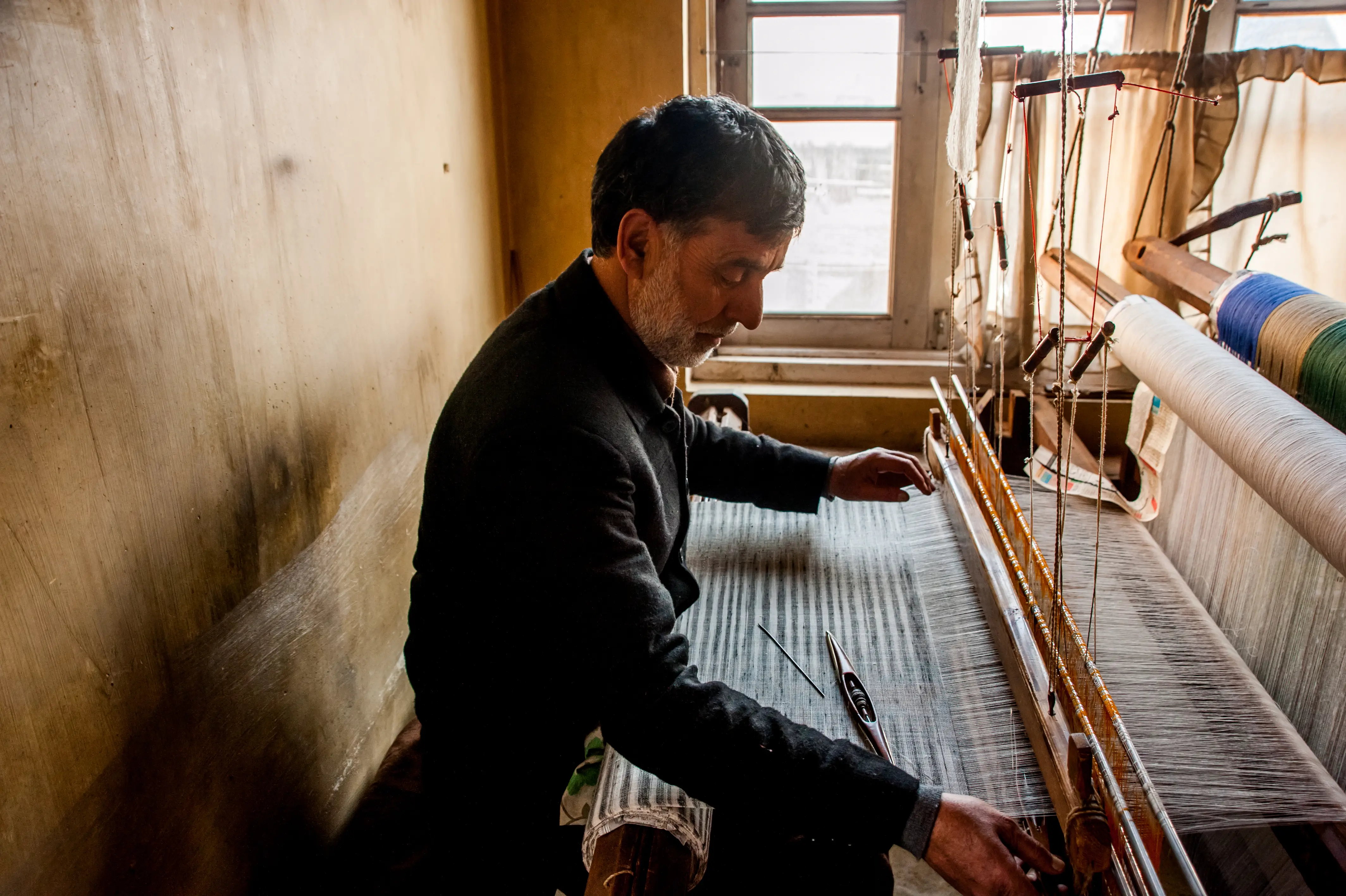 A weaver working by a window, surrounded by spools of coloured thread