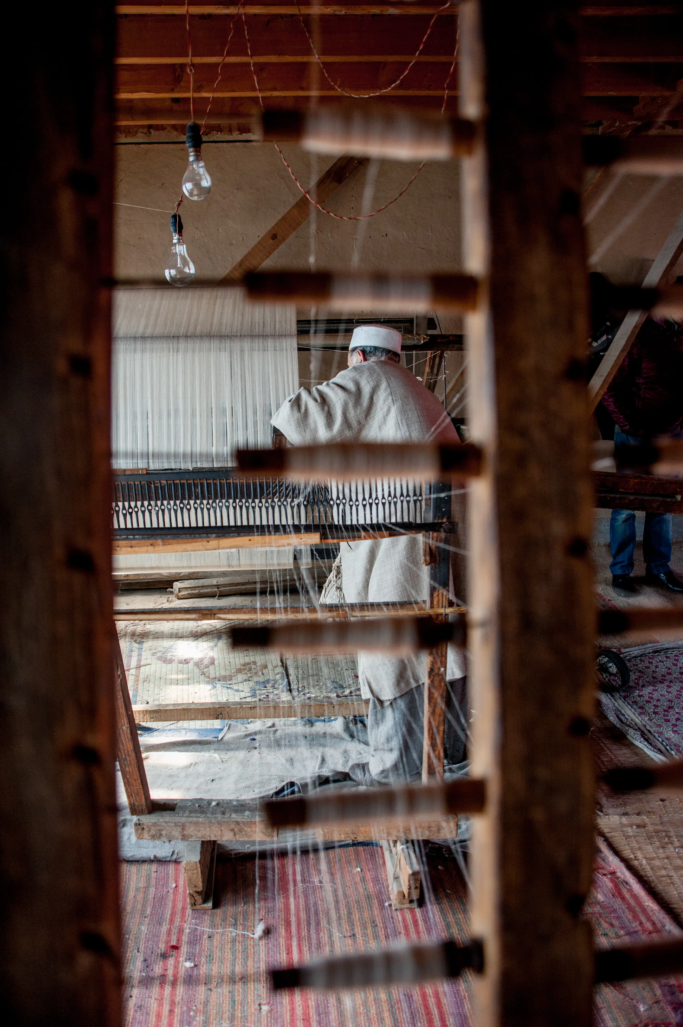 An elderly weaver seen from behind, working at a handloom framed by wooden lattice